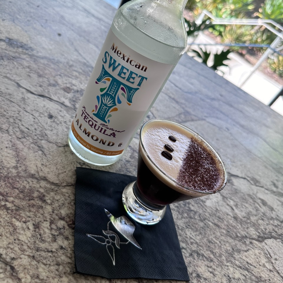 A glass of espresso martini topped with coffee beans next to a bottle of almond tequila.