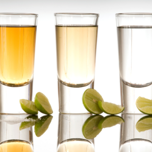 Three shot glasses of tequila in different colors with lime slices on a reflective surface.