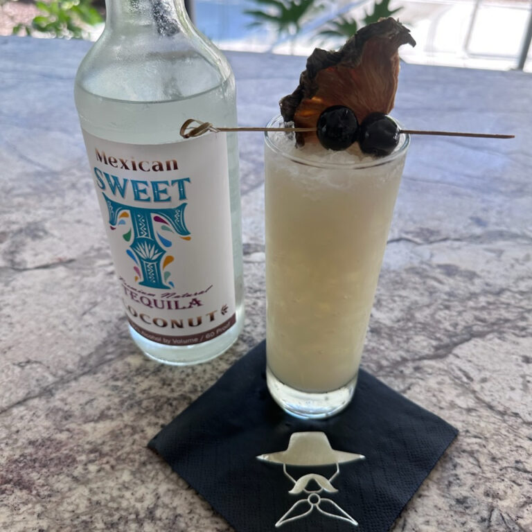 A glass of tequila pina colada garnished with dried fruit and berries, alongside a bottle of Mexican tequila.